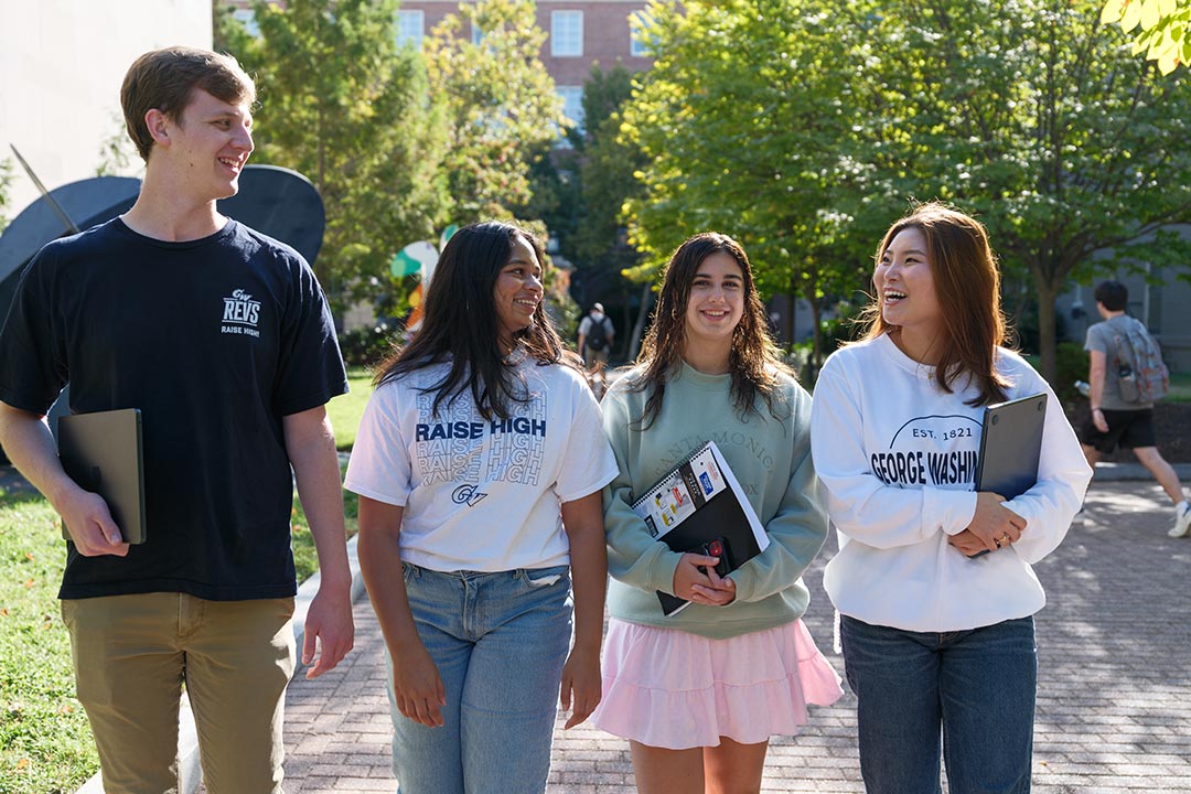 A group of four students in GW apparel walking around campus with green trees in the background